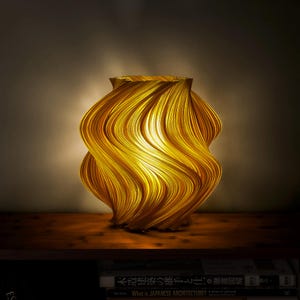 Puede incluir: Una lámpara de mesa con un exterior ondulado y acanalado de color amarillo dorado que brilla desde el interior. La lámpara presenta una forma orgánica compleja y en espiral con crestas verticales que crean un juego de luces y sombras.