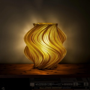 May include: A golden-yellow lamp with a layered, swirling design. The lamp is lit from within, creating a warm glow. It sits on a wooden shelf, with books visible below.