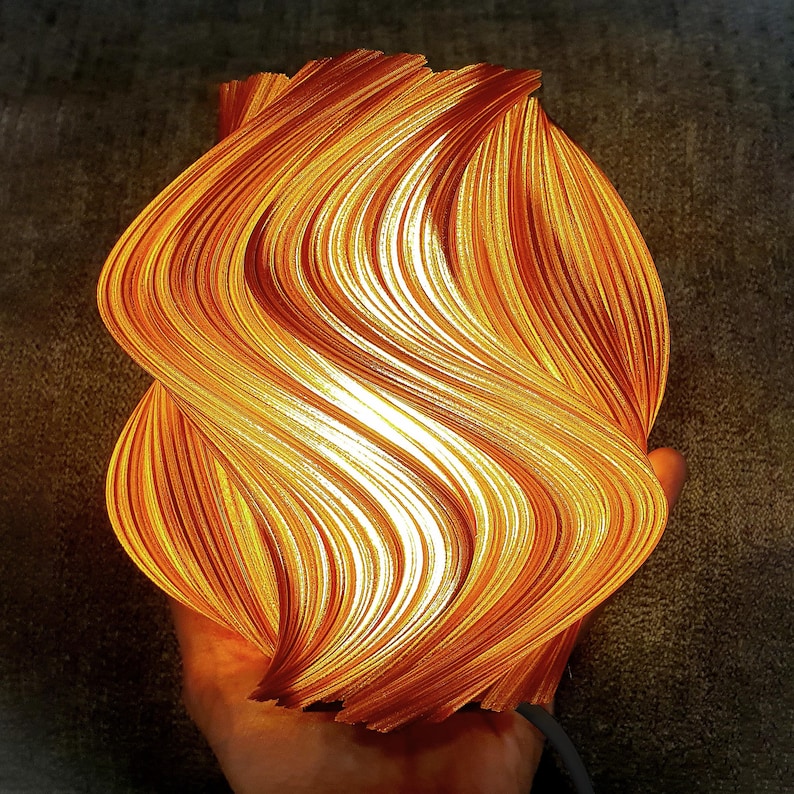 May include: A 3D printed lamp with a swirling, wave-like structure that glows with a warm orange and yellow light. The surface features intricate, vertical ridges that create a dynamic, flame-like appearance, emitting a soft, diffused illumination.