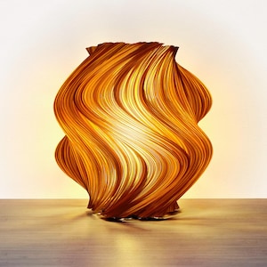 Lámpara decorativa con un diseño único y ondulado. La lámpara está hecha de finas tiras curvas de un material marrón dorado cálido, creando una apariencia texturizada en forma de ola. La fuente de luz interna emite un brillo suave y acogedor.