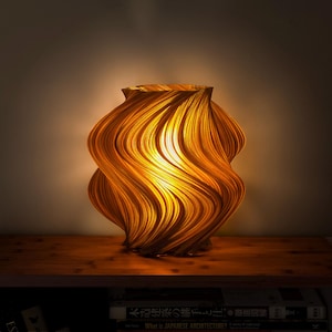 Puede incluir: Una lámpara de mesa con una estructura cilíndrica, en espiral y ondulada que emite una luz dorada y cálida. La lámpara presenta una superficie texturizada y acanalada que crea sombras profundas y reflejos brillantes, irradiando luz desde el centro.