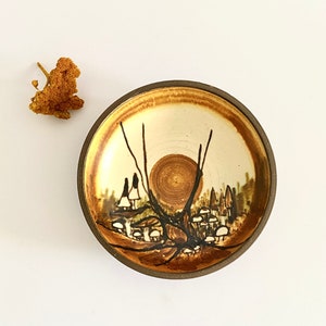 May include: A small, round ceramic bowl with a brown rim and a white interior. The bowl features a hand-painted design of a forest scene with trees, mushrooms, and a sun.