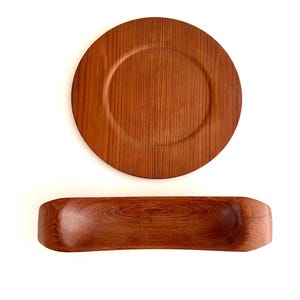 Puede incluir: Dos piezas de servicio de madera: un plato redondo y un cuenco rectangular. El plato tiene un borde elevado y el cuenco tiene bordes redondeados. Ambos están hechos de madera marrón cálido con vetas visibles.