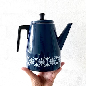 Puede incluir: Una cafetera de esmalte azul con un diseño floral blanco alrededor de la parte inferior. La cafetera tiene un asa y un pico negros.