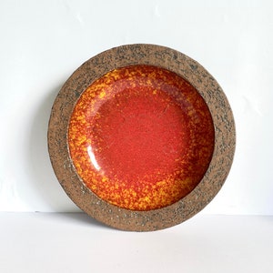 Puede incluir: Un plato redondo de cerámica con un borde marrón texturizado y un esmalte rojo y amarillo. El esmalte tiene un patrón moteado.