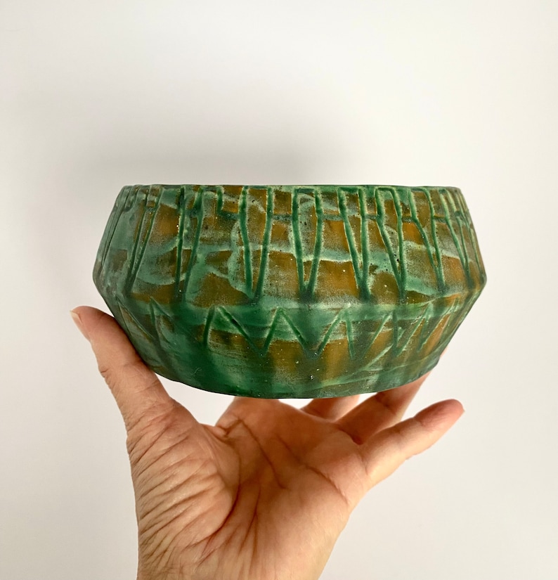Może przedstawiać: Zielono-brązowa doniczka ceramiczna z geometrycznym wzorem. Doniczka ma szeroką, zaokrągloną podstawę i lekko rozszerzający się rant.