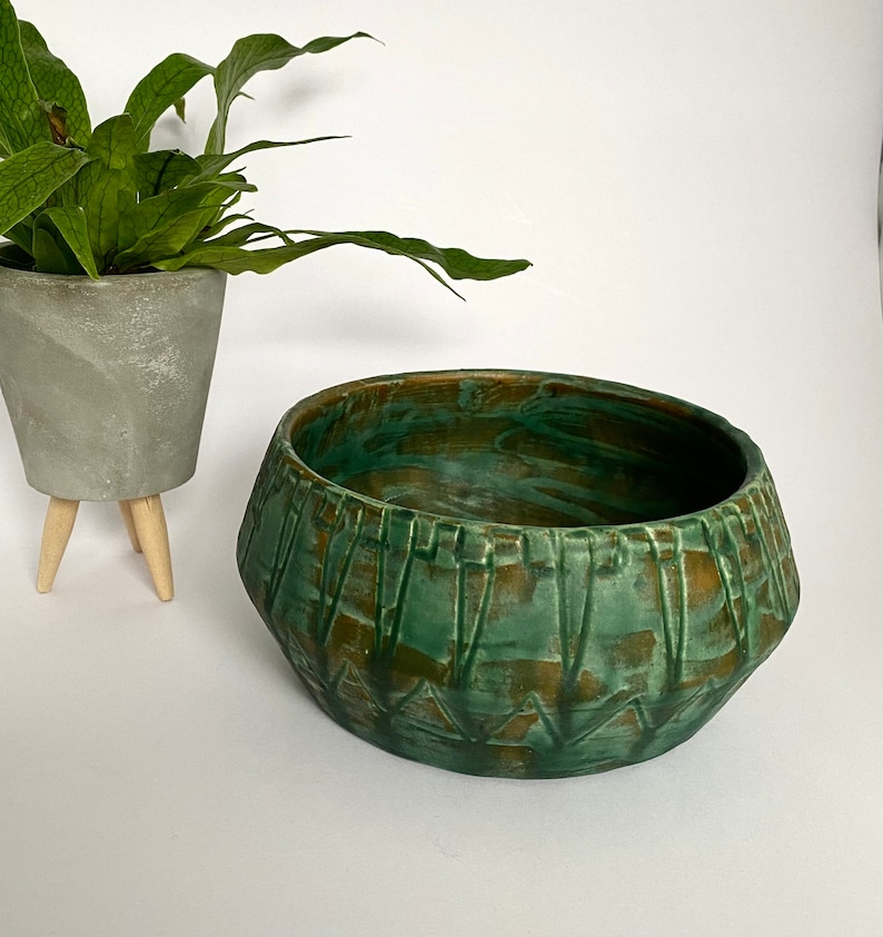 Może przedstawiać: Zielona, glazurowana doniczka ceramiczna z geometrycznym wzorem. Doniczka ma szeroki, płytki kształt misy i lekko rozszerzający się rant.