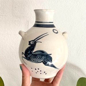 May include: Cream-colored ceramic vase with a dark blue antelope design. The vase has a rounded body, two small handles, and a cylindrical neck with dark blue stripes. The antelope is depicted in a running pose.