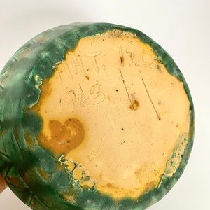 Może przedstawiać: Sp&oacute;d zielonego, szkliwionego wazonu ceramicznego z napisem "HAT. 1912" w kolorze brązowym.