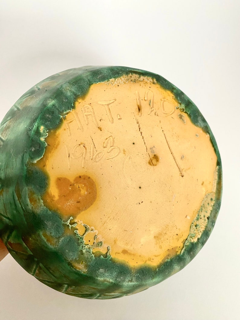 Może przedstawiać: Sp&oacute;d zielonego, szkliwionego wazonu ceramicznego z napisem "HAT. 1912" w kolorze brązowym.