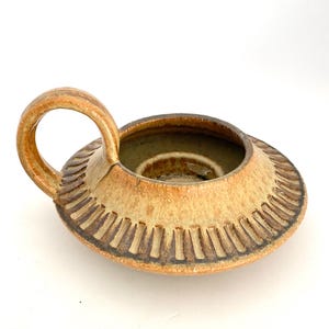 May include: A handmade ceramic candle holder with a circular design and a handle. The candle holder is tan with brown accents and a textured pattern around the rim. The interior is a darker shade of brown.