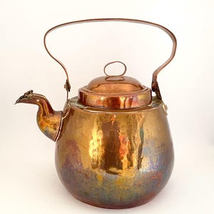 Könnte beinhalten: Ein Vintage-Kupferkessel mit gebogenem Griff und kleinem Deckel. Der Kessel hat einen abgerundeten Körper und einen Ausguss mit vogelartigem Design. Das Kupfer hat eine polierte, reflektierende Oberfläche mit schillernden Farbtönen.