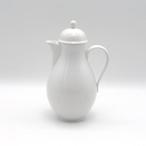 Puede incluir: Una cafetera de cerámica blanca con tapa y asa. La cafetera tiene un cuerpo redondeado, un pico y un asa curvada. La tapa está rematada con un pequeño pomo. La cafetera está sobre un fondo blanco.