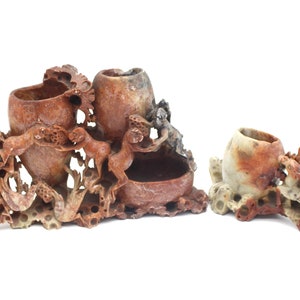 May include: A set of three carved stone sculptures. The largest piece has three cylindrical openings and is decorated with figures and foliage. The other two pieces have one opening each and are also decorated with carved details. The stone is a mix of brown, tan, and gray.