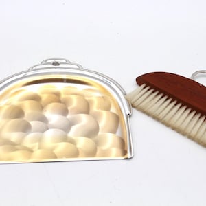 Puede incluir: Un recogedor plateado con interior texturizado y un cepillo con mango de madera y cerdas blancas. El cepillo tiene un anillo de metal para colgarlo. El conjunto está diseñado para la limpieza.