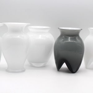 May include: A collection of six glass vases in various shapes and sizes. The vases are primarily white, with one dark gray vase. The vases have a smooth, glossy finish and are arranged against a white background.