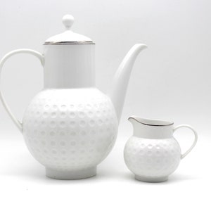 Peut inclure: Un ensemble de cafetière et de pot à crème en céramique blanche. La cafetière a un corps haut et cylindrique avec une poignée incurvée et un bec verseur. Le pot à crème est petit et rond avec une poignée. Les deux pièces ont un motif texturé et alvéolé et une garniture argentée.