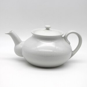 Peut inclure: Une théière en céramique blanche avec un corps arrondi, un bec verseur incurvé et une anse en boucle. La théière a un couvercle avec un petit bouton. Le design est simple et élégant, idéal pour servir le thé. Sur fond blanc.