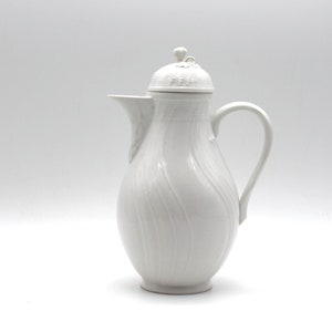Könnte beinhalten: Weiße Porzellan-Kaffeekanne mit Deckel und einem wirbelnden Design. Der Deckel hat einen kleinen Knopf oben.