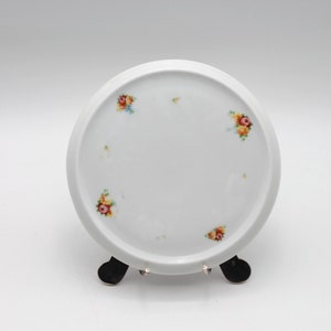 May include: A white, round ceramic plate with a slightly raised rim. The plate is decorated with small floral clusters in shades of red, yellow, and blue. The plate is displayed on a black metal stand.