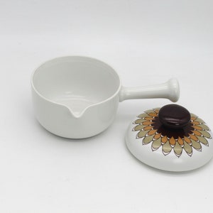 Puede incluir: Cacerola de cerámica blanca con asa y boquilla. La tapa presenta un diseño floral en tonos marrón, naranja y beige. Un artículo de cocina vintage.