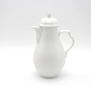 Könnte beinhalten: Weiße Porzellan-Kaffeekanne mit Deckel und Henkel. Die Kanne hat einen bauchigen Körper und einen schlanken Ausguss. Die Oberfläche ist mit einem zarten Blumenmuster verziert. Der Griff hat ein dekoratives, geschwungenes Design.