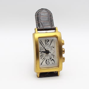 Puede incluir: Un reloj de pulsera rectangular dorado con esfera blanca y números negros. El reloj tiene una correa de cuero marrón y tres botones en el lateral. La esfera muestra el nombre de la marca y otros detalles.