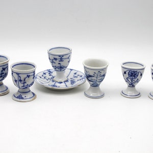 May include: A set of seven white porcelain egg cups with blue floral designs and gold trim. The egg cups are of varying designs, with one resting on a matching saucer. A classic and elegant addition to any breakfast table.