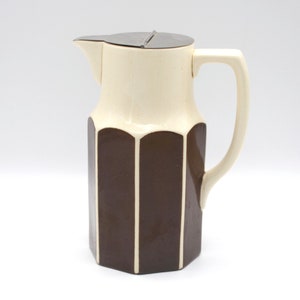 Op de afbeelding: Een vintage crème- en bruinkleurige keramische kan met een deksel. De kan heeft een geometrisch ontwerp met verticale bruine panelen en een crème-kleurig bovendeel. Het heeft een gebogen handvat en een schenktuit.