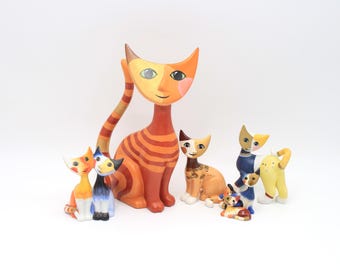Può includere: Una collezione di sette statuette di gatti di varie dimensioni e colori. Il gatto più grande è arancione a strisce, mentre i più piccoli presentano motivi in blu, giallo, marrone e bianco. Ogni statuetta ha un volto stilizzato con occhi dipinti.
