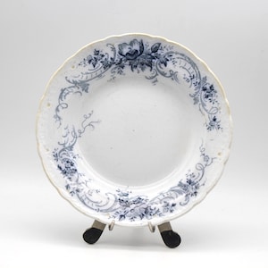 May include: White ceramic plate with a scalloped edge and a blue floral design. The plate features a detailed pattern of flowers and vines around the rim. The plate is displayed on a small metal stand.