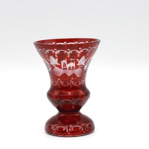 Puede incluir: Un jarrón de cristal rojo rubí con la parte superior acampanada y una base de pedestal. El jarrón presenta diseños blancos grabados de follaje y un paisaje. El jarrón mide unos 15 cm de alto.