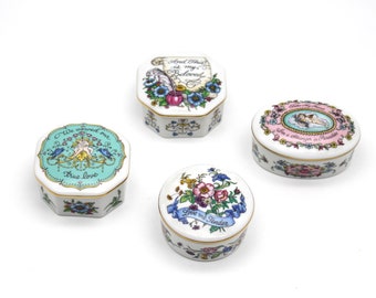 May include: Four decorative porcelain trinket boxes with floral and romantic designs. The boxes are white with gold trim and feature various shapes, including octagonal and round. Each box has a unique design with text, flowers, and cherubs.