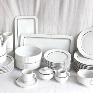 Op de afbeelding: Een complete witte porseleinen servies set met een getextureerd, schorsachtig patroon. De set bevat borden, kommen, kopjes, schotels, een theepot, serveerschalen en een melkkan. Elk stuk heeft een dunne, metallic gouden rand.