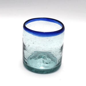 Puede incluir: Un vaso de vidrio transparente soplado a mano con un borde azul vibrante. El vidrio tiene una superficie ligeramente texturizada y una forma redondeada. El vaso es ideal para servir agua, zumo o cócteles. El vaso mide aproximadamente 10 cm de alto.