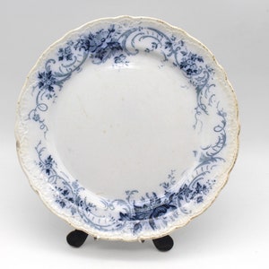 May include: White ceramic plate with a scalloped edge and a blue floral design. The plate has a gold trim and is displayed on a black stand. The floral pattern includes flowers and swirling vines.