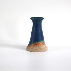 May include: A handmade ceramic vase with a distinctive design. The vase has a dark blue top that fades into a lighter blue, with an orange band and a textured beige base. The vase has a flared shape.