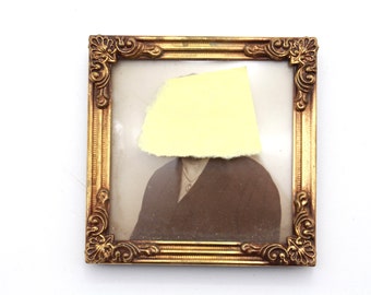 Puede incluir: Una fotografía vintage enmarcada con un marco dorado ornamentado. La fotografía presenta a una persona que lleva una parte superior de color oscuro y un collar. Un trozo de papel amarillo cubre la cara de la persona en la fotografía.