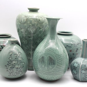 May include: A collection of six celadon ceramic vases in varying shapes and sizes, featuring light green glaze with intricate inlaid designs including floral patterns, willow trees, and cranes, all displayed together against a plain white background.