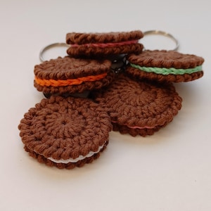 May include: Four crocheted cookie keychain charms with different colored fillings. The cookies are brown with white frosting and a colored filling. The keychains are attached to silver key rings.