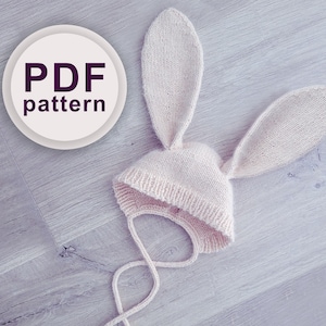 Può includere: Un cappello di coniglio lavorato a maglia con orecchie lunghe e una stringa di cravatta. Il cappello è di colore crema chiaro. L'immagine include il testo "PDF pattern".