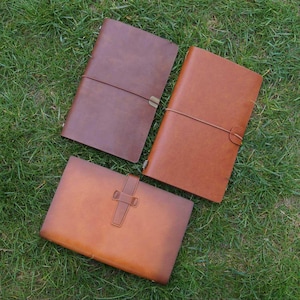 May include: Three brown leather journals are arranged on green grass. The journals feature a classic design with either a strap or elastic closure. The leather appears smooth and well-crafted, suggesting a high-quality product, suitable for writing.