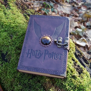 Może przedstawiać: Brązowy skórzany dziennik z złotym zapięciem i kamieniem tygrysiego oka. Dziennik ma wygrawerowane na przodzie słowa "Harry Potter".