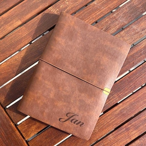 May include: A brown leather-bound journal with a gold clasp and the name "Jan" engraved on the front.