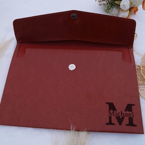 May include: A brown leather envelope with a silver snap closure and a personalized monogram of the letter "M" with the name "Michael" underneath.