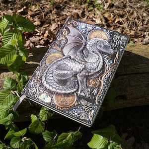 Könnte beinhalten: Ein silbernes und golden geprägtes Journal mit einem Drachen-Design. Der Drache ist silber mit goldenen Akzenten und von floralen und geometrischen Mustern umgeben. Das Journal hat einen strukturierten Einband und ein silbernes Lesezeichenband.