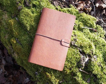 May include: A brown leather bound journal with a string closure resting on a bed of green moss.
