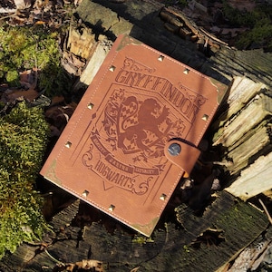 Może przedstawiać: Brązowy skórzany dziennik z złotymi akcentami i wygrawerowanymi na okładce słowami "Gryffindor" i "Hogwarts". Dziennik zawiera również herb Gryffindoru z lwem i napisami "Odwaga, Śmiałość, Rycerskość".