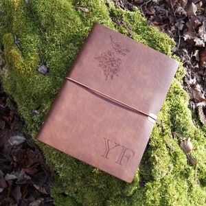 May include: A brown leather journal with a floral design and the initials "YF" embossed on the cover. The journal is lying on a bed of green moss.