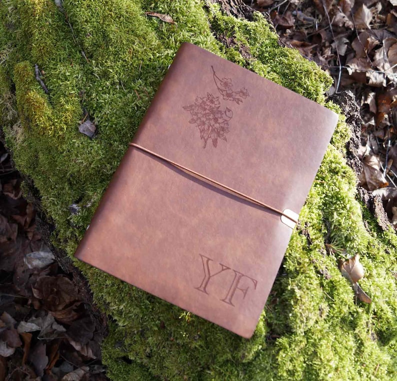 May include: A brown leather journal with a floral design and the initials "YF" embossed on the cover. The journal is lying on a bed of green moss.